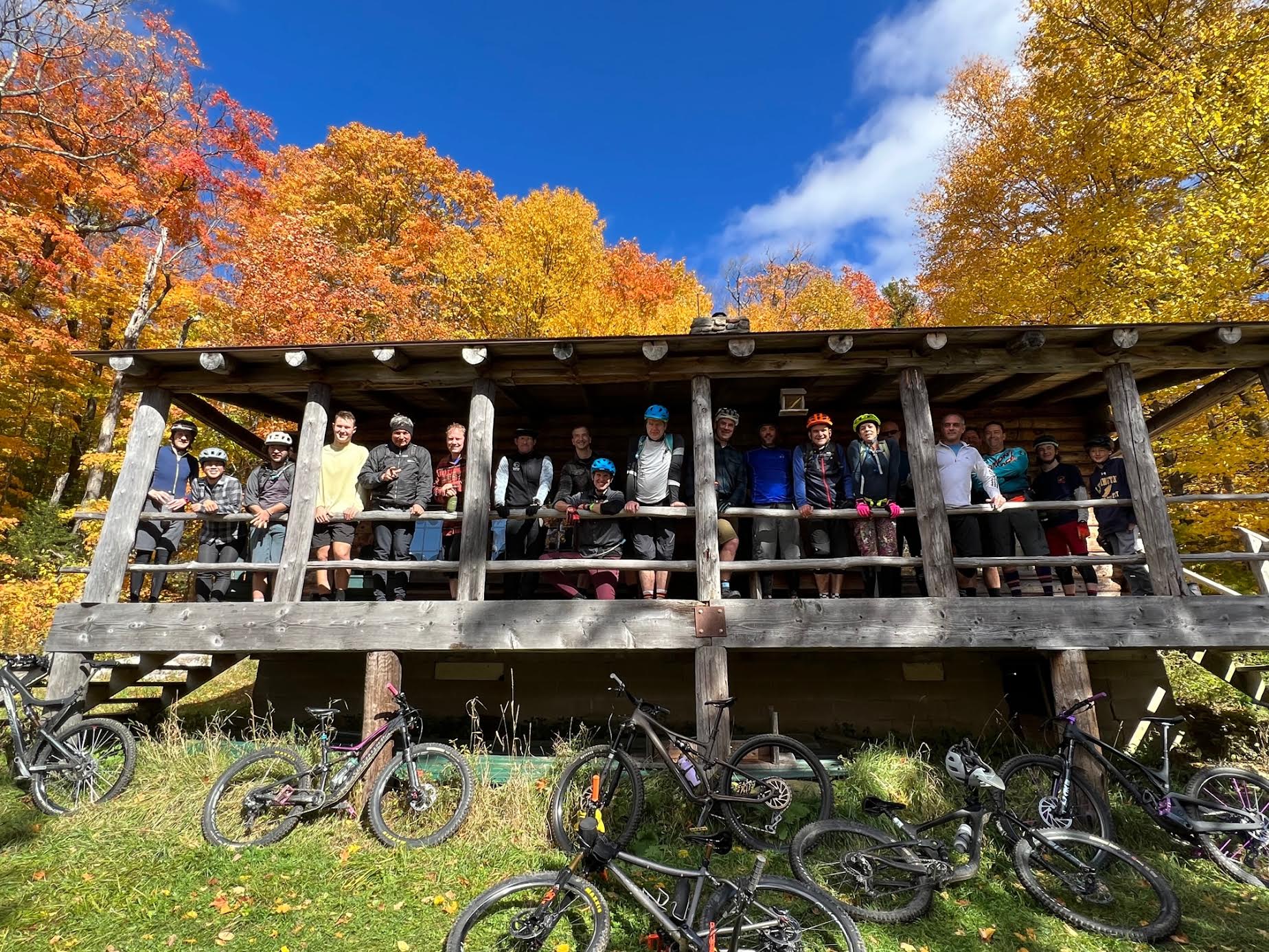 A group of people stand on the porch of a log cabin wearing bike helmets and smiling.